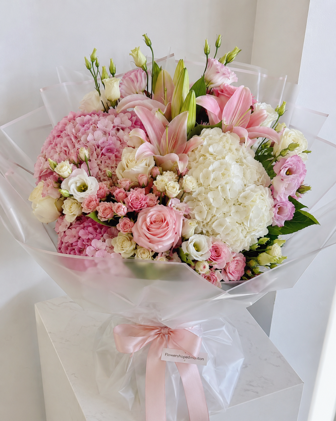 A beautifully arranged floral bouquet featuring pink hydrangeas, lilies, white and pink roses, and white flowers, wrapped in clear cellophane with a light pink ribbon. Edmonton flowers delivery 