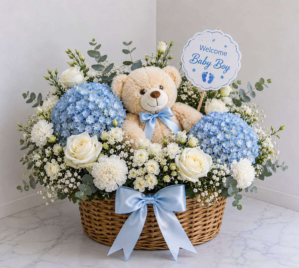 A baby boy flower basket arrangement in a wicker basket, featuring light blue hydrangeas, white roses, baby's breath, eucalyptus, a beige teddy bear with a blue ribbon, and a round sign that reads 'Welcome Baby Boy'.